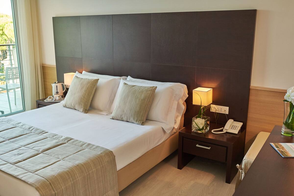 A hotel room with a large bed, a bedside table with a lamp, a phone, and a small vase with flowers.