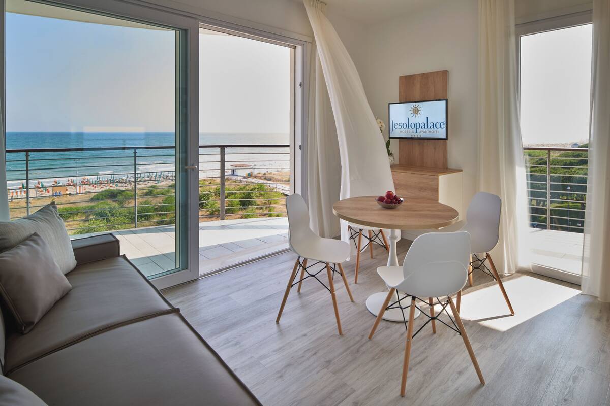 A modern living room with a large balcony, a wooden dining table with chairs, and a TV mounted on the wall.