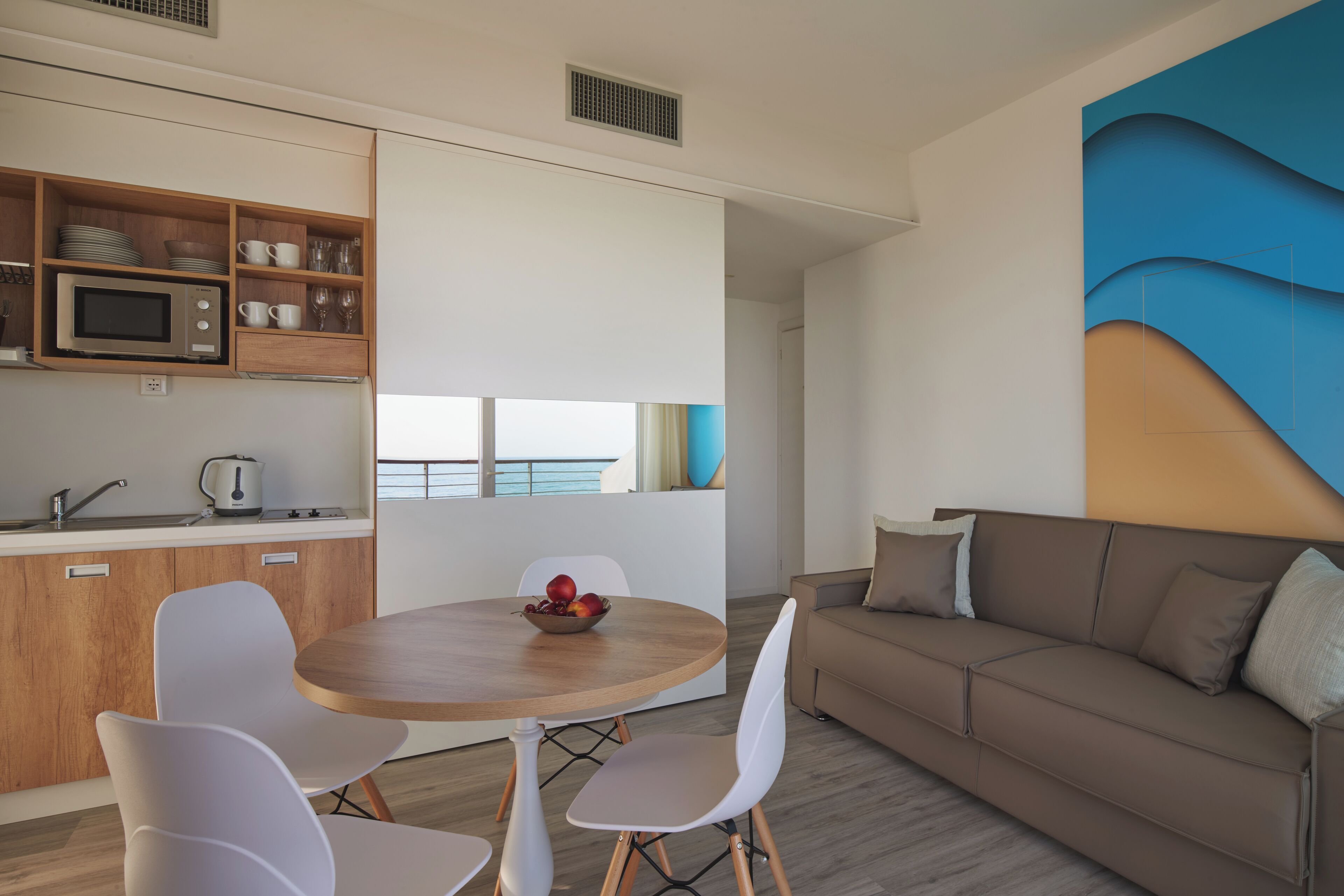 A modern kitchen with a microwave, sink, and wooden cabinets. There is a dining area with a round table and white chairs. A brown sofa is against the wall, and a large abstract painting is on the wall.