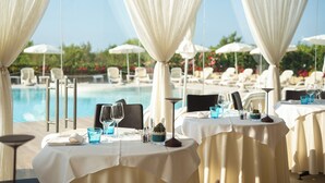 Ein Essbereich am Pool mit Tischen für Zwei, weißen Tischdecken und Blick auf Pool und Liegestühle.