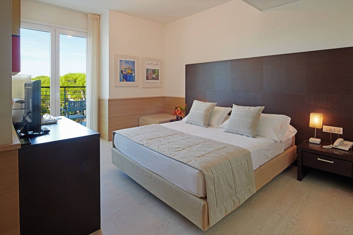 A hotel room with a large bed, a desk with a computer, a bedside lamp, and a view of the outdoors through a window.