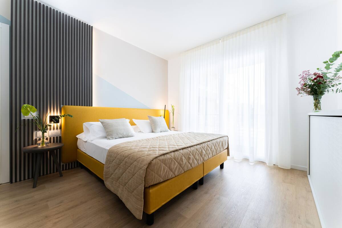 A modern bedroom with a yellow bed, white bedding, a bedside table with a lamp, and a wall-mounted panel.