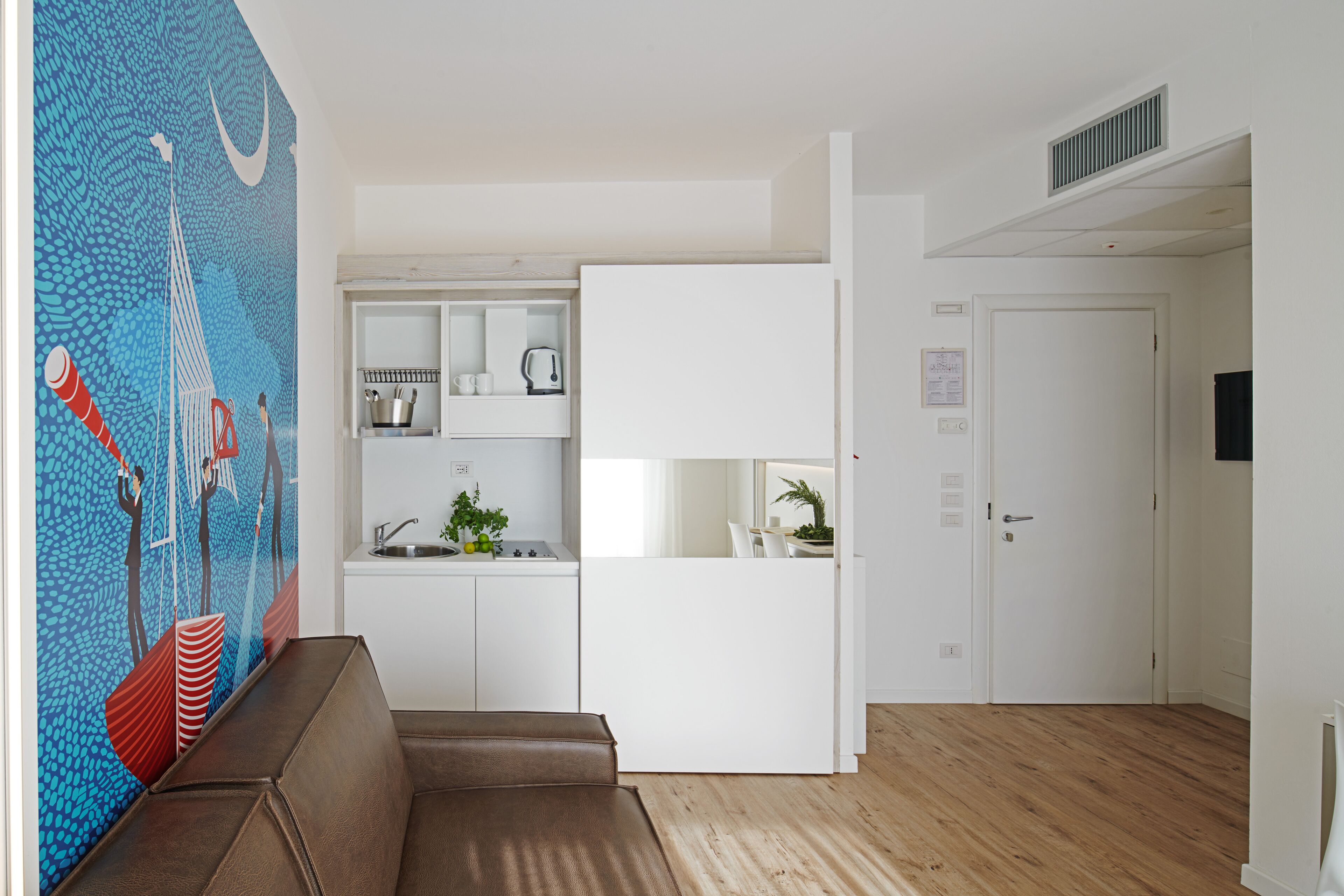 A modern kitchen with white cabinets, a sink, and a countertop. There is a brown sofa in the living area and a door leading to another room.