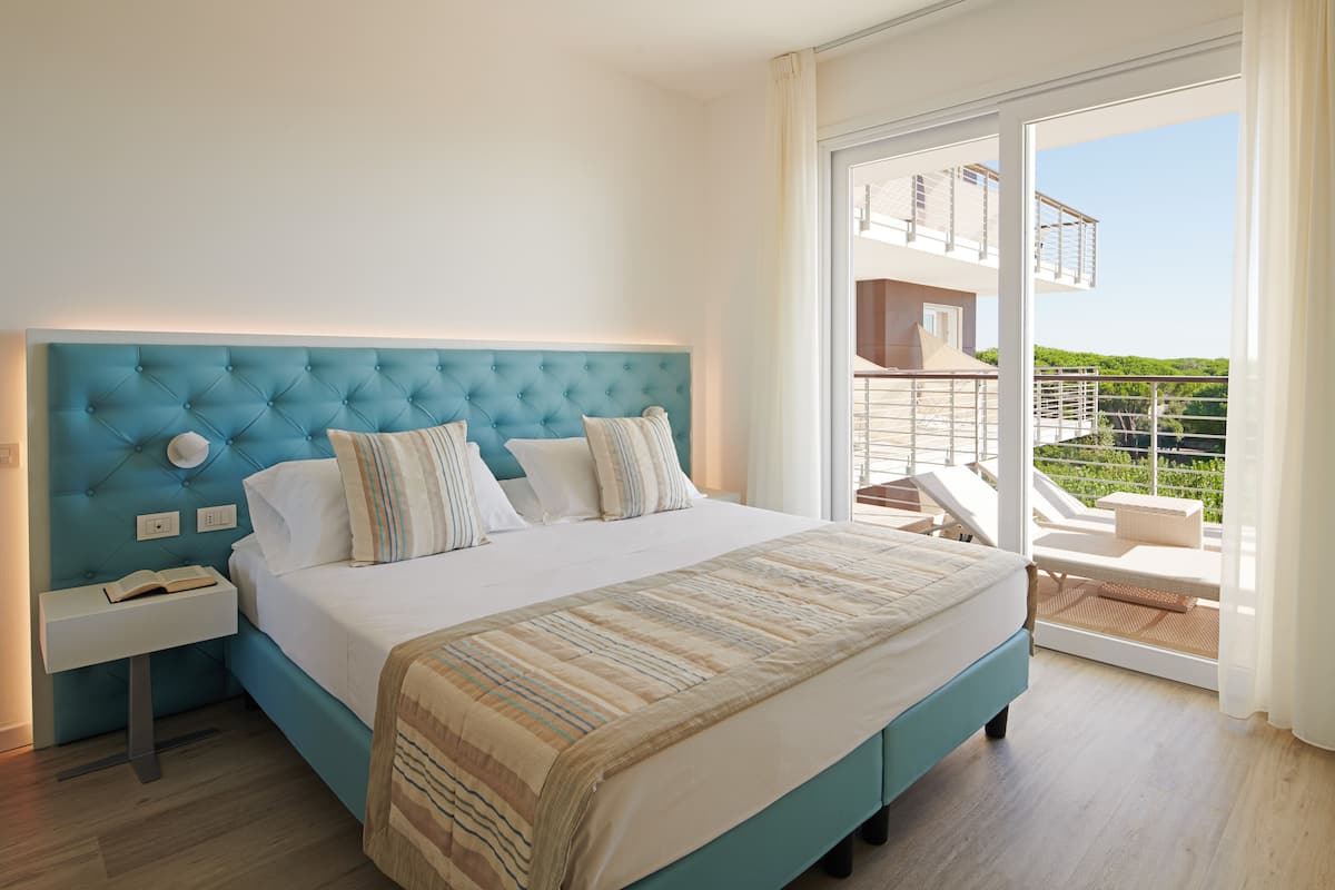 A bedroom with a large bed, a nightstand, and a view of a balcony with outdoor furniture.