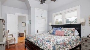 A bedroom with a floral bedspread, a wooden floor, a black headboard, a nightstand, and a window with blinds.