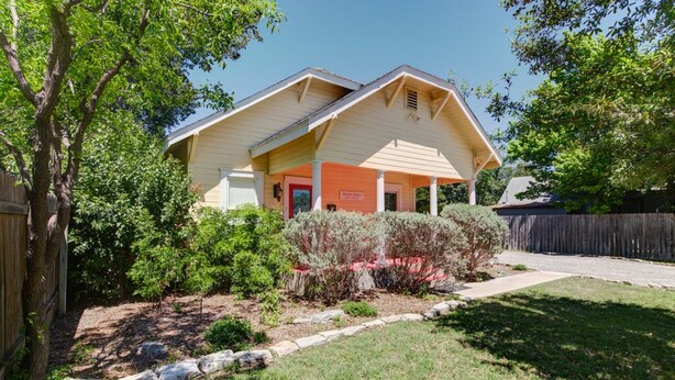 A single-story house with a sloped roof, a front porch, and a small garden area with various shrubs and a tree.