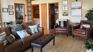 Un vaste salon comprenant un canapé en cuir marron, un fauteuil en cuir marron et un banc. Le plafond est orné de poutres en bois, on y trouve également une échelle en bois et une porte qui donne sur une autre pièce.