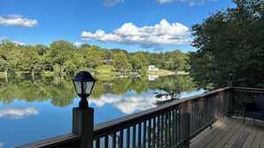 Une terrasse en bois avec un lampadaire, d’où l’on peut admirer un lac paisible dont les eaux reflètent les arbres et une maison au loin.