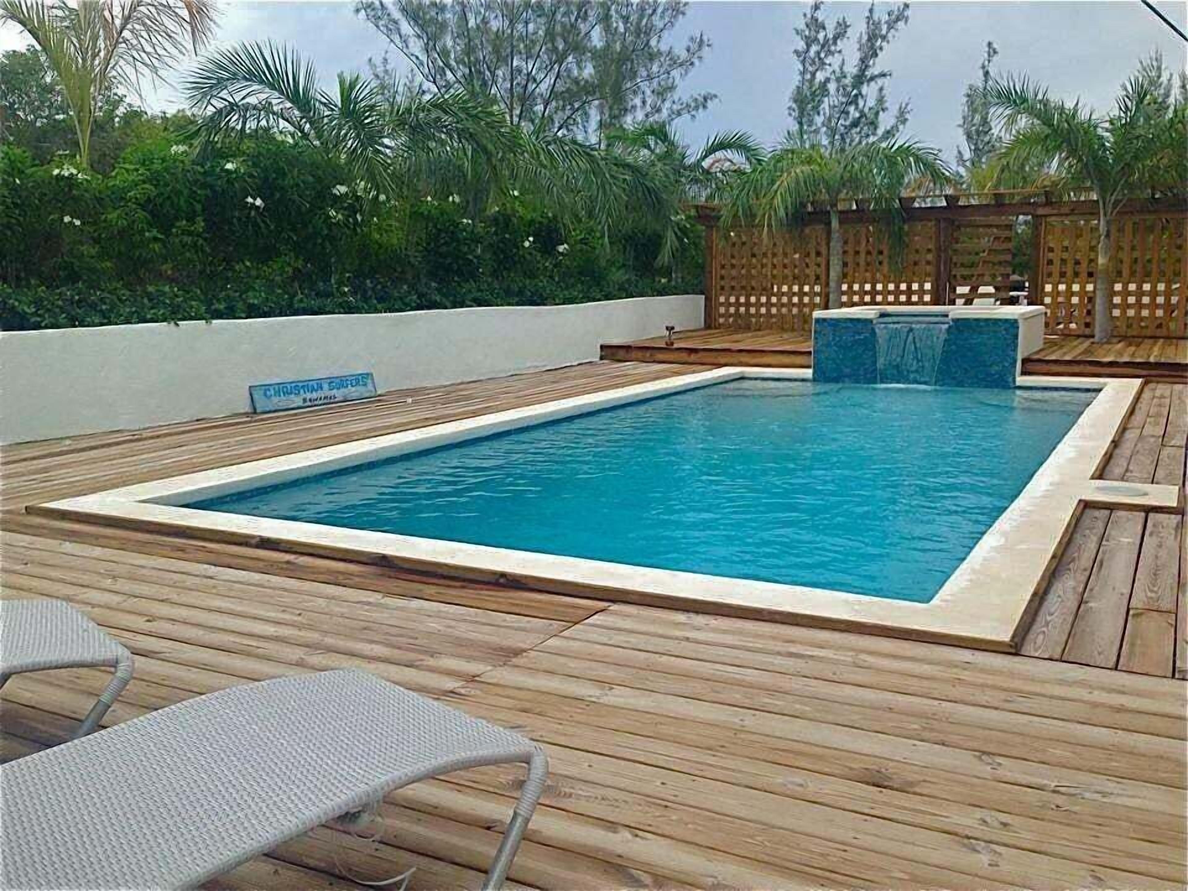 Piscina retangular com um deck de madeira ao redor, uma cerca de madeira ao fundo e uma placa que diz 'CHRISTIAN SURFES'.
