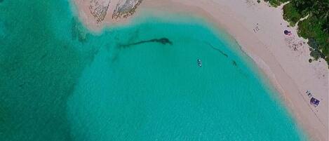 Ovenfra ses en strand med krystalklart turkis vand, sandet bredde og vegetation.