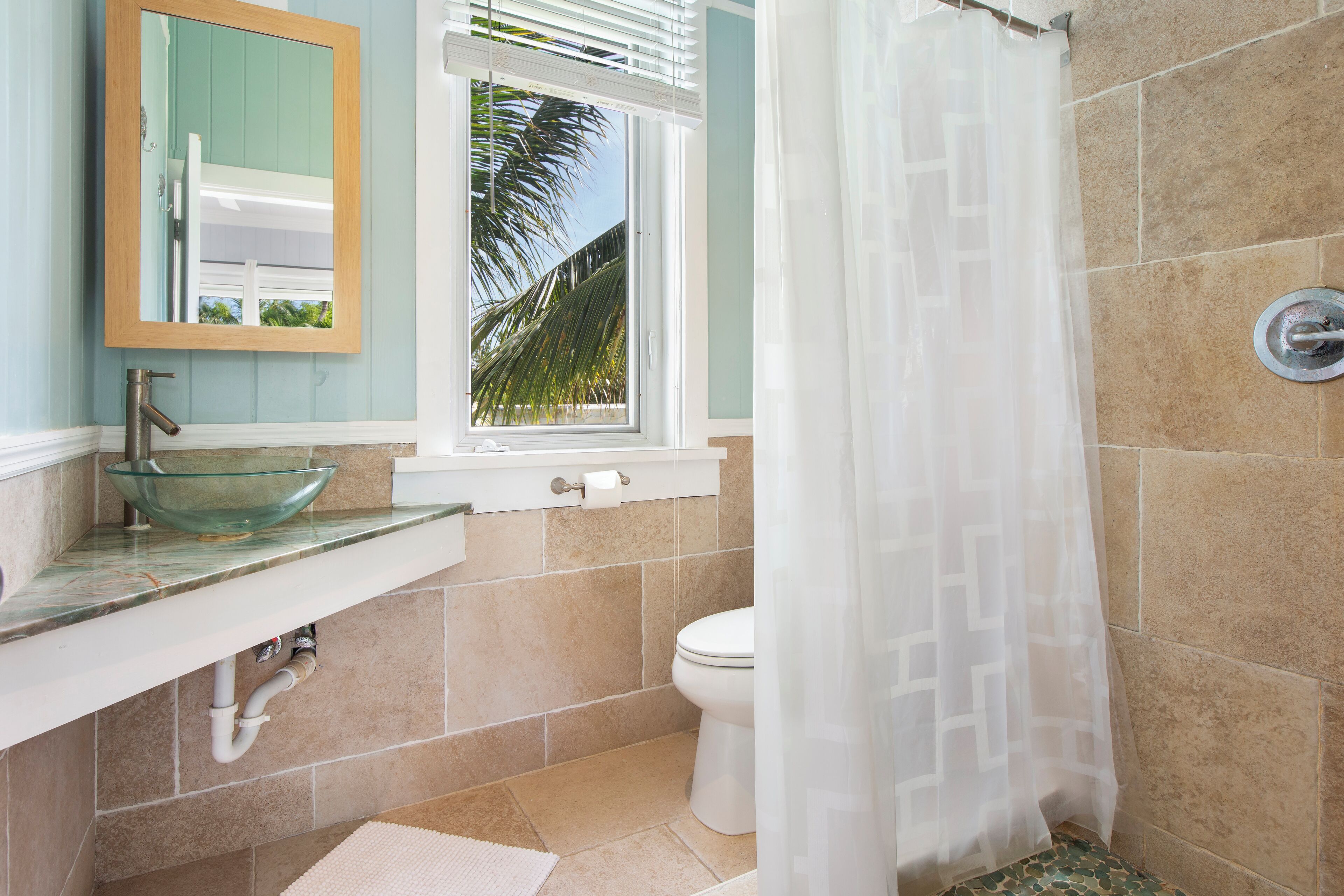 Une salle de bain avec un lavabo en forme de bol en verre, des toilettes et une fenêtre donnant sur des palmiers.