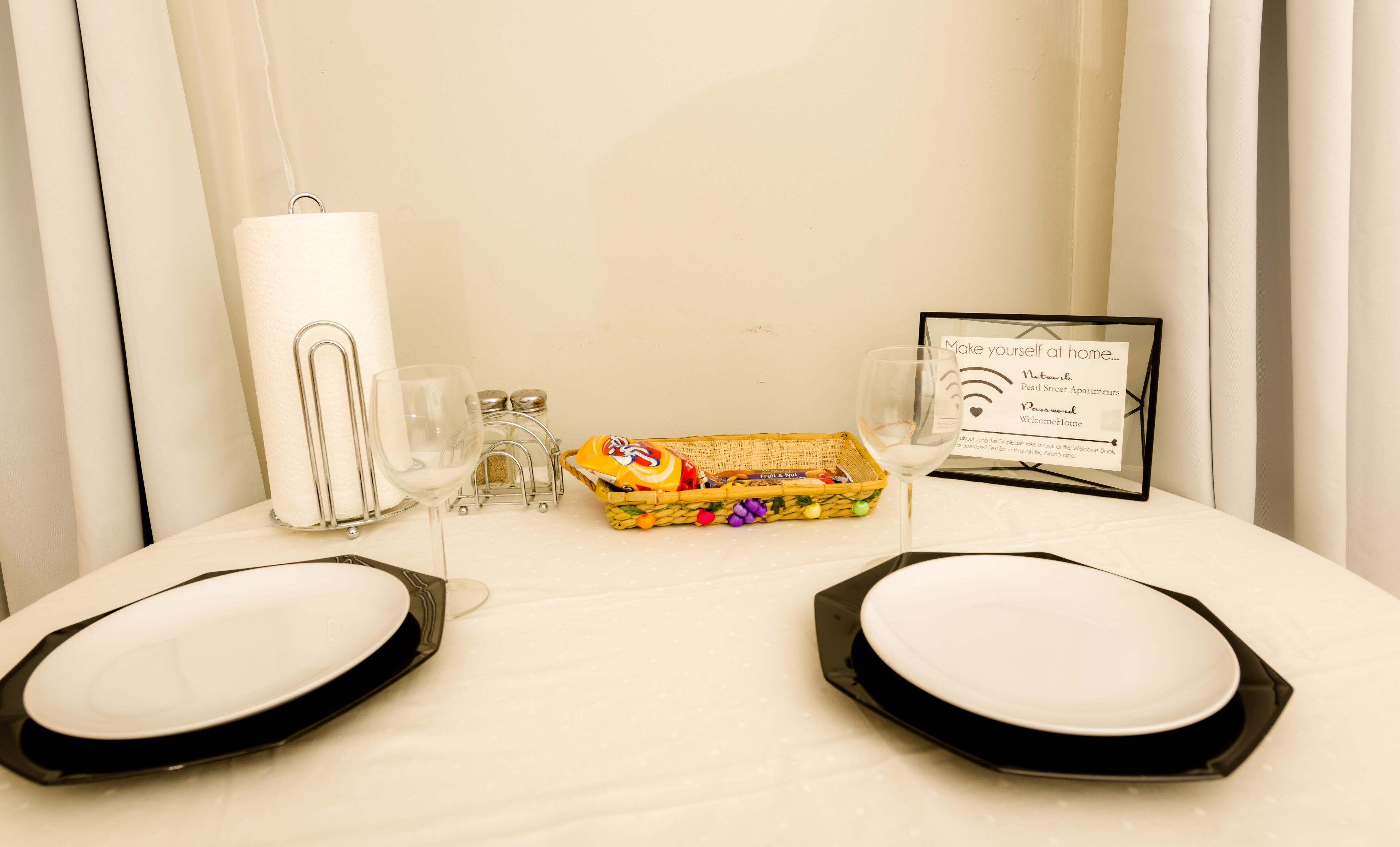 Hotelkamer met een eettafel, inclusief een mand fruit, een glas water, zout- en pepervaatjes, en een welkomstbord.