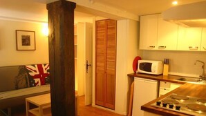 A compact kitchen with a microwave, stove, and wooden cabinets. A wooden pillar and a door with a Union Jack flag are visible. - RENT CHARMING STUDIO (Cauterets)