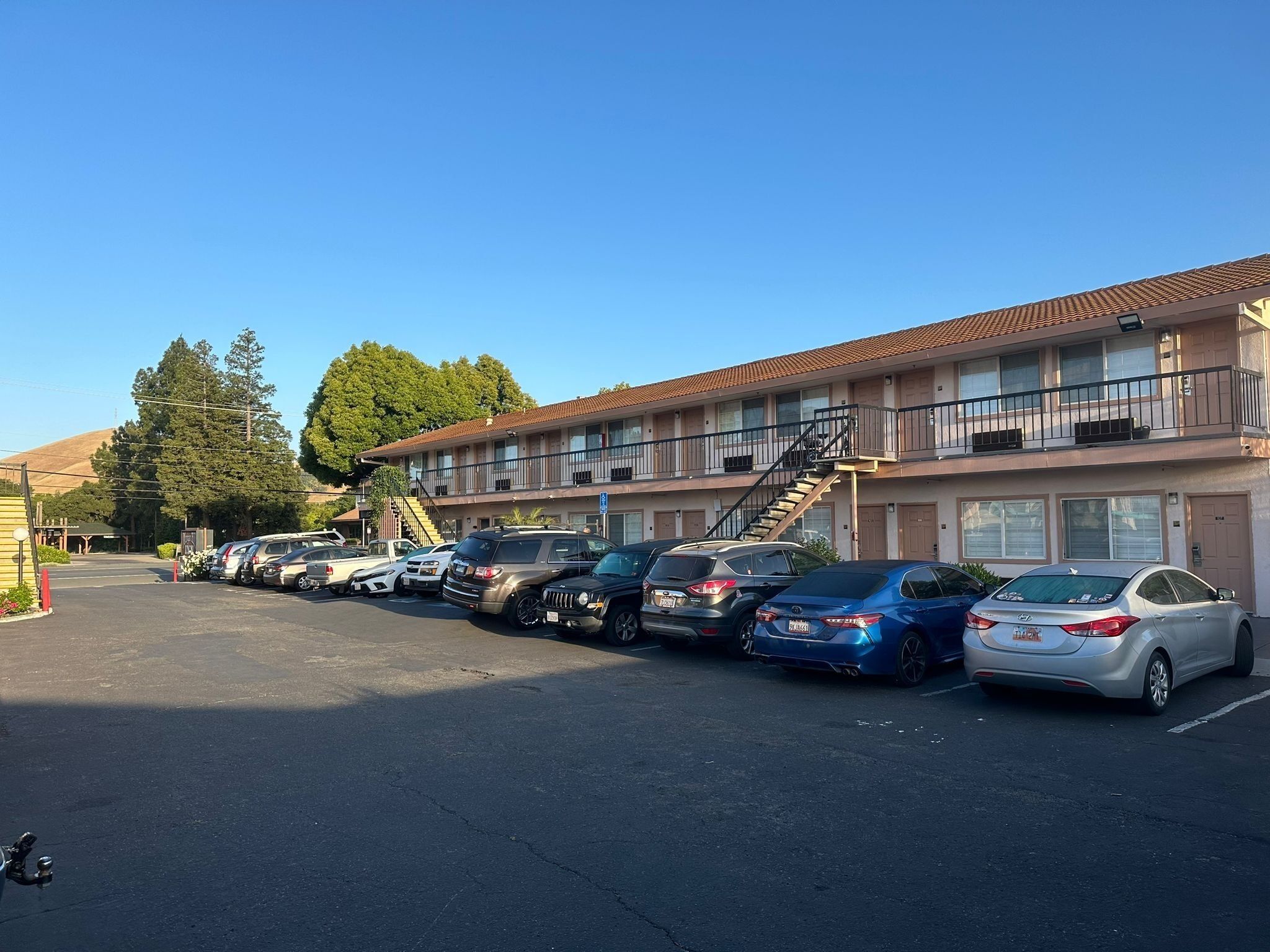 A motel with a row of parked cars in front, a staircase leading to the upper floor, and a clear blue sky.