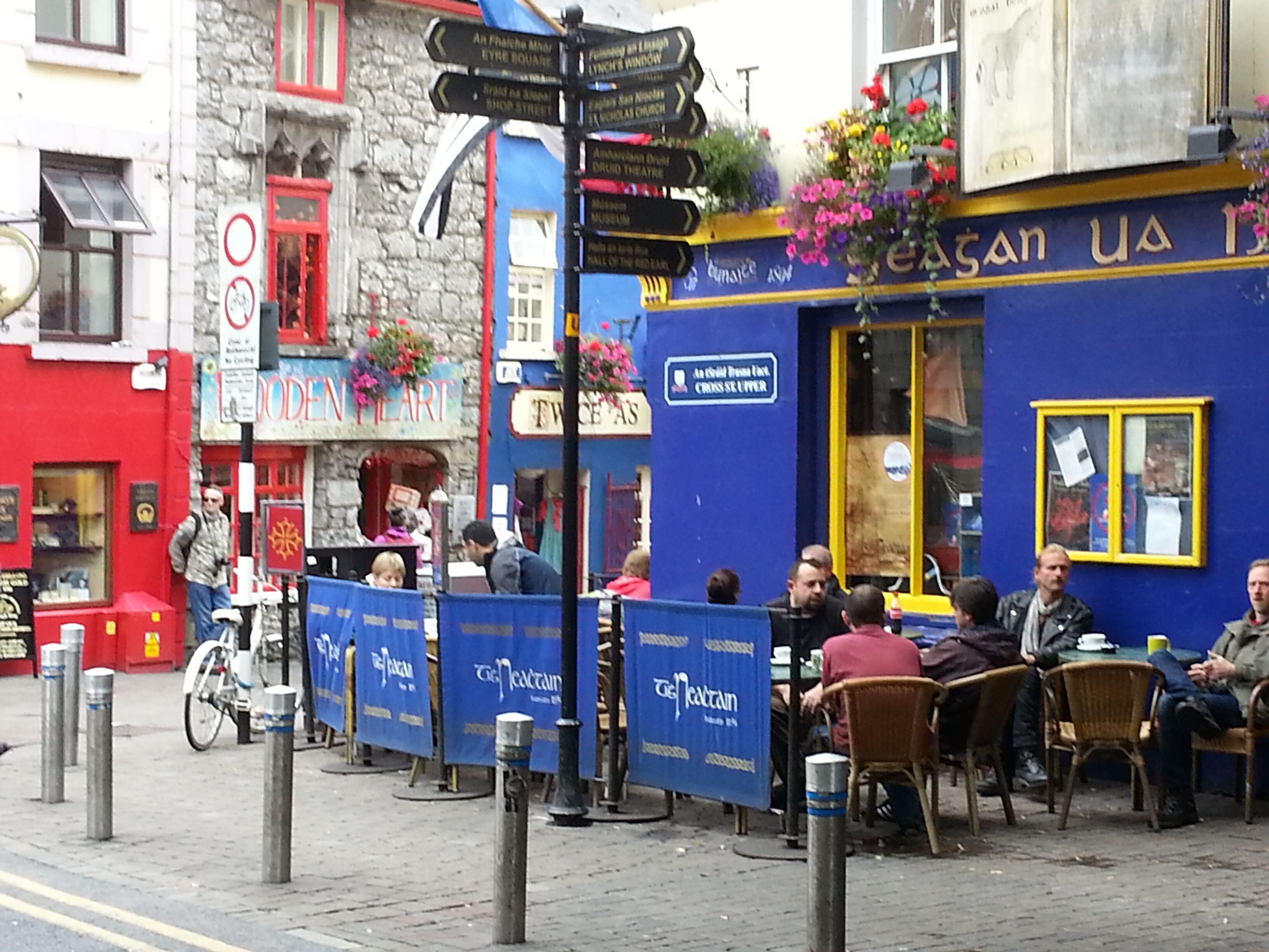 A street scene with a blue building, a signpost with directions, and people sitting outside.