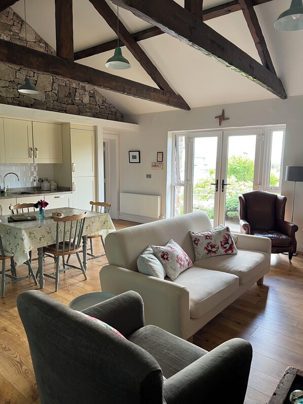 Un salon chaleureux avec un canapé, des fauteuils, une table à manger et un coin cuisine aux murs en pierre et aux poutres en bois.