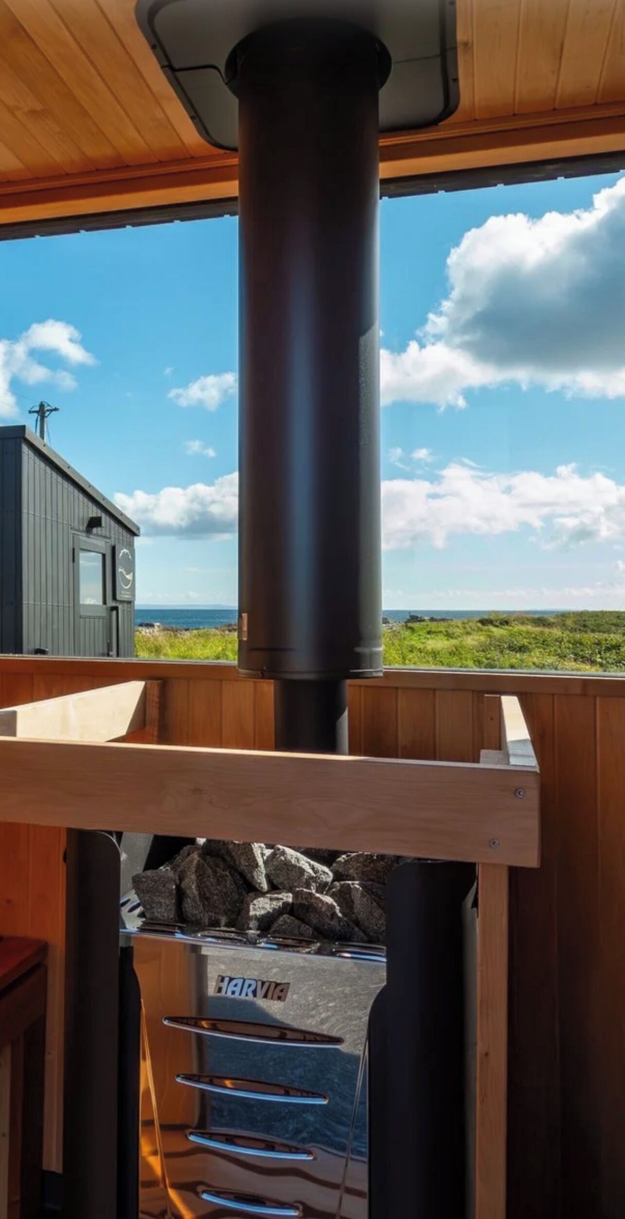 Une terrasse en bois avec une cheminée noire, un barbecue Harvia et une vue sur la mer et le ciel.