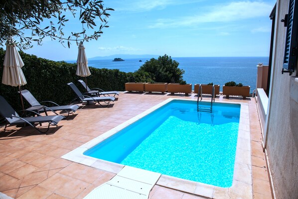 A rectangular swimming pool with a clear blue water, surrounded by lounge chairs and umbrellas, with a view of the sea and distant islands.
