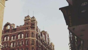 Un bâtiment historique à étages, doté d’arcades et de fenêtres en arc de cercle, situé dans un quartier urbain animé, entouré d’autres immeubles.