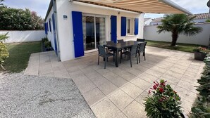 Un patio con mesa y sillas, una zona de estar cubierta y un pequeño jardín con plantas y una palmera.