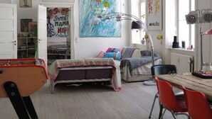 A bedroom with a bed, a desk, and a chair. There is a window with curtains, a radiator, and a shelf with books and a lamp.
