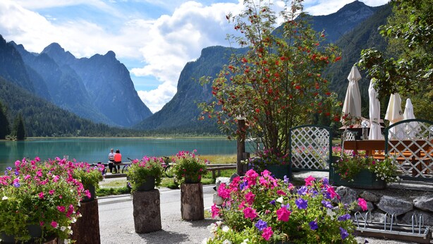 Un patio junto al lago con flores en macetas, un banco de madera y vistas a las montañas.
