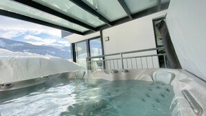 Un jacuzzi con recinto de vidrio, barandilla metálica y vista a una montaña nevada.