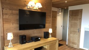 A wooden-paneled room with a flat-screen TV mounted above a wooden console table, flanked by two wall-mounted lamps. - SKIS ON FOOTS-WIFI- GARAGE (optional) -NICE VIEW-2 ROOMS (Les Allues)