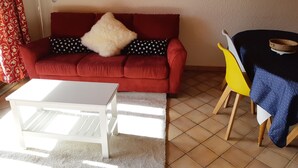 Ein Wohnzimmer mit einem roten Sofa, einem weißen Couchtisch, einem gelben Sessel und einem Tisch mit blauer Tischdecke.