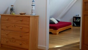 A room with a wooden dresser, a small table with a jar and a bowl, a bed with a red blanket, and a bookshelf in the background.