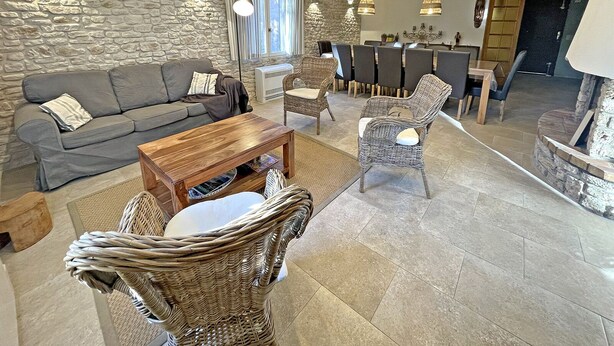 Een gezellige zithoek met een stenen muur, een grijze bank, rieten stoelen, een houten salontafel en een eethoek met stoelen en een tafel.