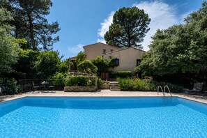 A swimming pool with a clear blue surface, surrounded by greenery and a building with a sloped roof.