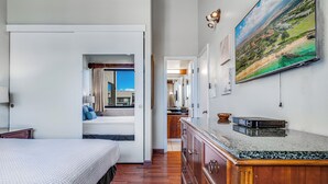 A hotel room with a bed, a wooden dresser, a painting, and a view of the beach. - Large 3 Bedroom Condo Across From Beach! Pool & Hot Tub Access! Ocean View! (Kihei)