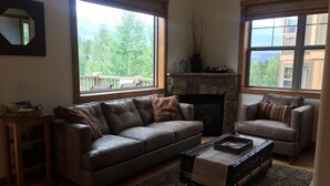 A living room with a stone fireplace, two leather sofas, a coffee table, and large windows with a view of greenery. - Breckenridge Ski Area Views from 4 Bedroom, 4 Bath Home (Breckenridge)
