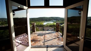 An open door leading to a balcony with a stone floor, a view of a river, and a forested hill in the distance.