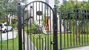 Une entrée fermée par un portail, avec une plaque d’immatriculation, un chemin menant à un bâtiment et une voiture blanche garée à proximité.