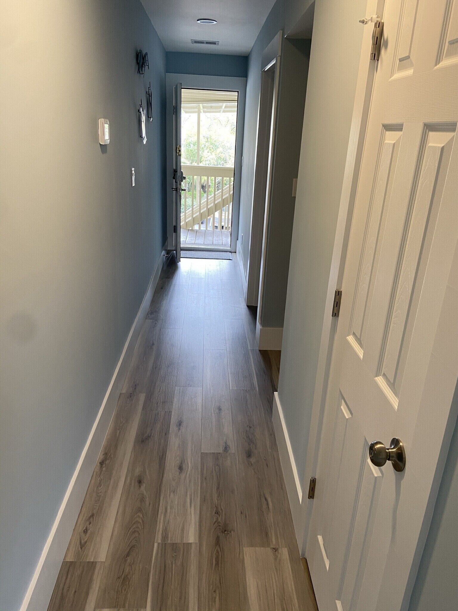 A hallway with a wooden floor, a door on the right, and a view of a balcony with a railing.