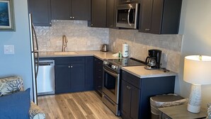Eine kompakte Küche mit dunklen Schränken, einem Edelstahlkühlschrank, einer Mikrowelle und einer Kaffeemaschine. Zwei Sofas mit gemusterten Bezügen und ein Holzboden.