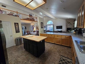 Una cocina con una isla central, armarios de madera, un comedor con sillas, una escalera y un televisor montado en la pared.