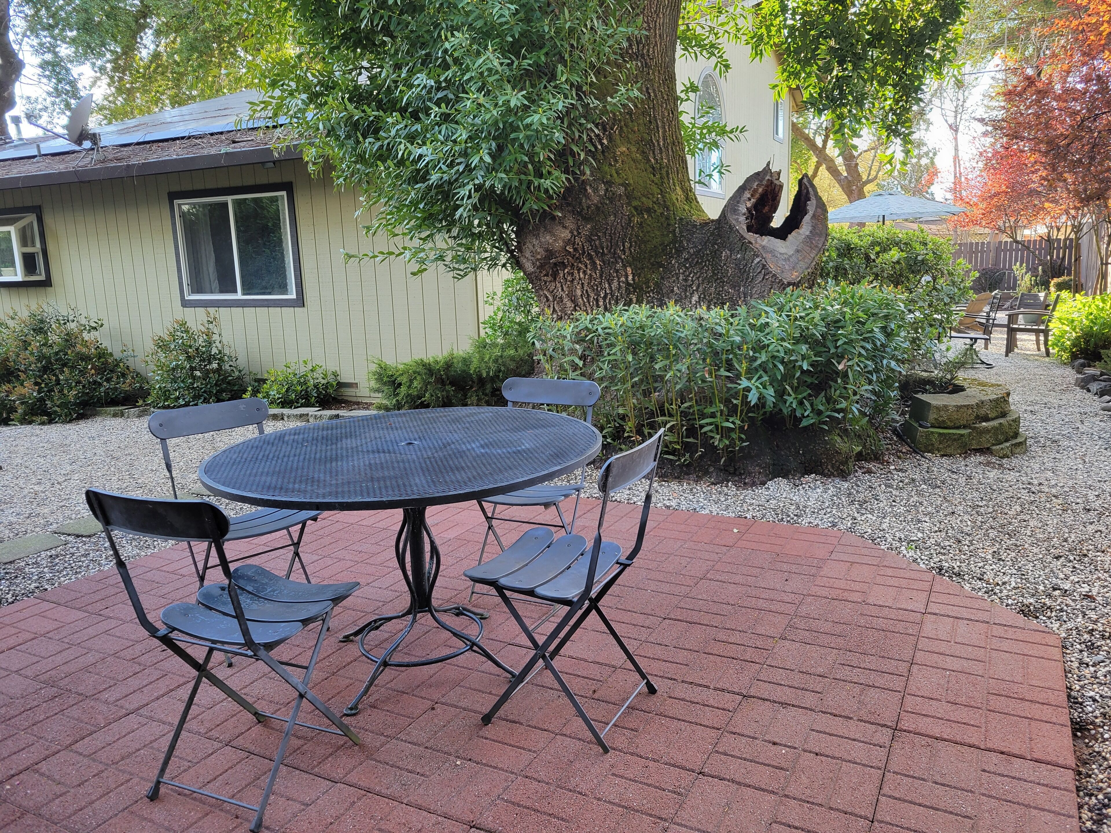 A patio with a round table and four chairs, surrounded by gravel and brick flooring, with a house and trees in the background.