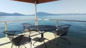 Un espace extérieur abrité, avec une table et des chaises, offrant une vue sur une étendue d’eau calme et des montagnes lointaines.