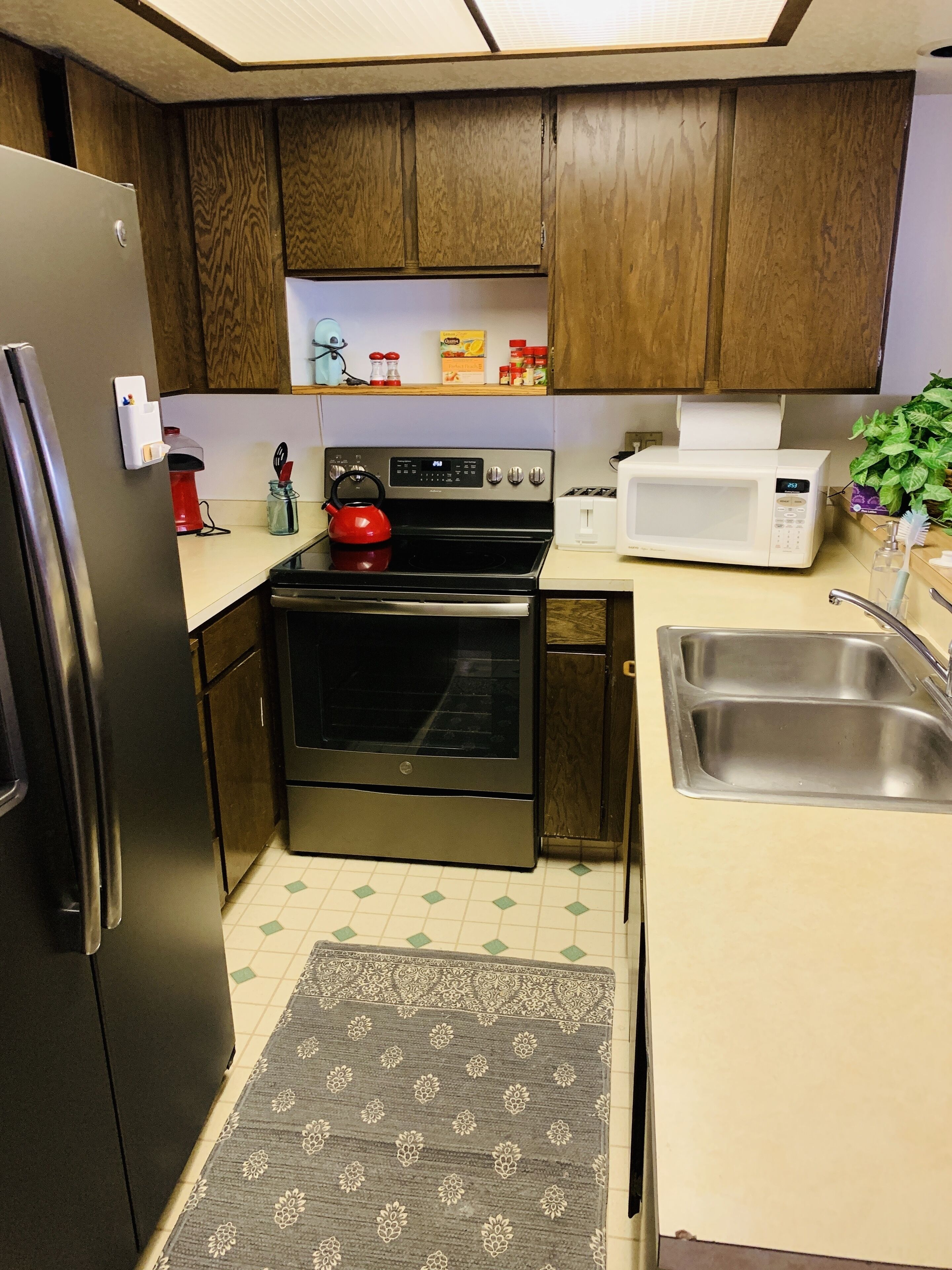 A compact kitchen with wooden cabinets, a stainless steel refrigerator, a red kettle, a microwave, and a sink.