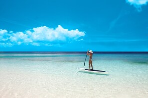 En person står på paddleboard på en klar strand med turkis vand og en strålende blå himmel.