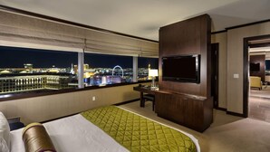 A hotel room with a large bed, a desk with a chair, a flat-screen TV mounted on the wall, and a view of the cityscape at night.