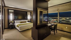 A hotel room with a large bed, a desk with a chair, a dresser, and a large window offering a city view at night.