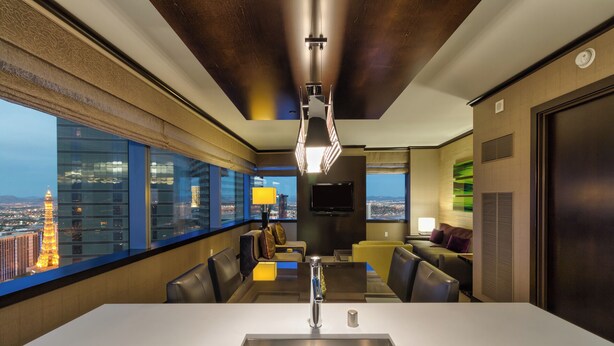 A modern hotel room with a large window offering a city view, a dining area with a white table and chairs, and a flat-screen TV mounted on the wall.