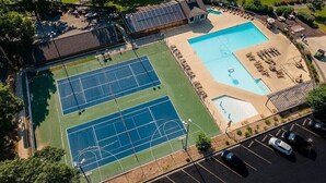 An aerial view of a property with a tennis court, a swimming pool, and a covered area with seating.