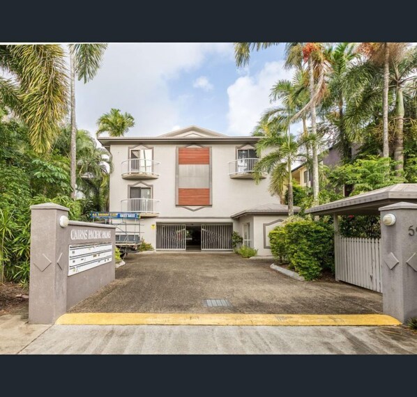 A two-story building with a gated entrance, surrounded by palm trees and greenery.