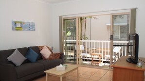 Una sala de estar con un sofá, una mesa de centro, un televisor y un ventanal corredizo que da a un espacio exterior con una mesa y sillas.