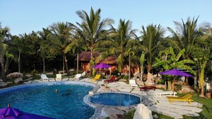 Un área de piscina con sillones de descanso y sombrillas, rodeada de palmeras y un pequeño edificio.